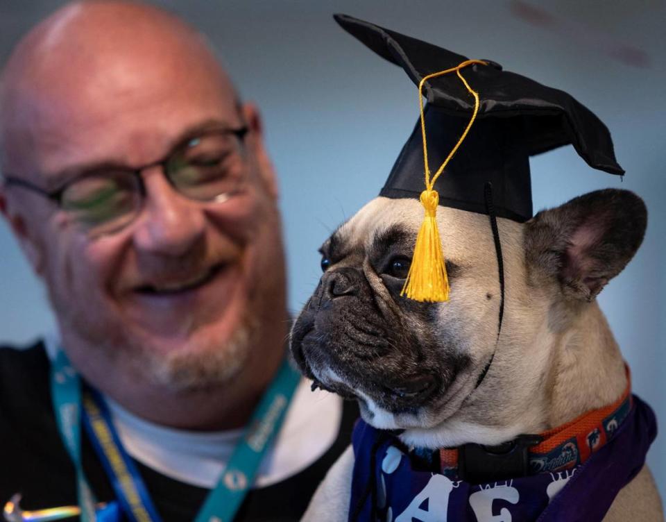 Eduardo García Rubio, a la izquierda, quien enseña producción televisiva en Hialeah Gardens High School, muestra a su perro, Bacardí, un bulldog francés que lleva una gorra de graduación durante un evento para promover la salud mental en la escuela secundaria. Hialeah Gardens, Florida - 30 de mayo de 2023 -