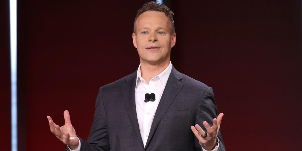 chris licht stands and holds his hands out in front of him, he is wearing a gray suit with a white collared shirt in front of a mostly red background