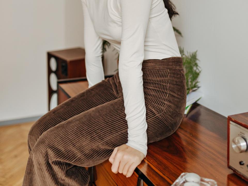 woman sitting on a side table wearing brown corduroys, a cream turtleneck, and a brown cap