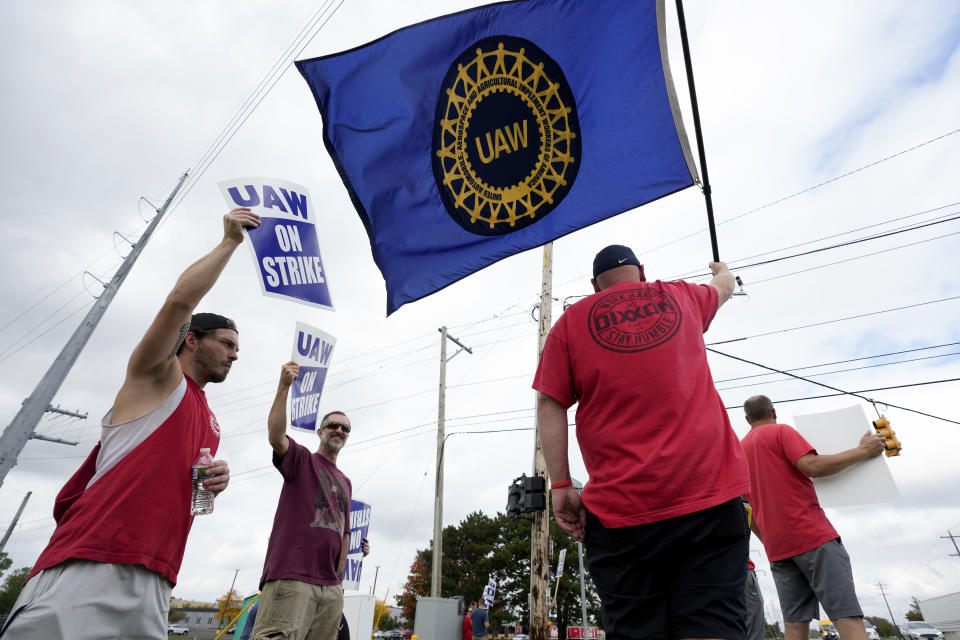 United Auto Workers members hold picket signs and a flag near a General Motors Assembly Plant in Delta Township, Mich., Friday, Sept. 29, 2023. The United Auto Workers union expanded its two-week strikes against Detroit automakers Friday, adding 7,000 workers at a Ford plant in Chicago and a General Motors assembly factory near Lansing, Michigan. (AP Photo/Paul Sancya)