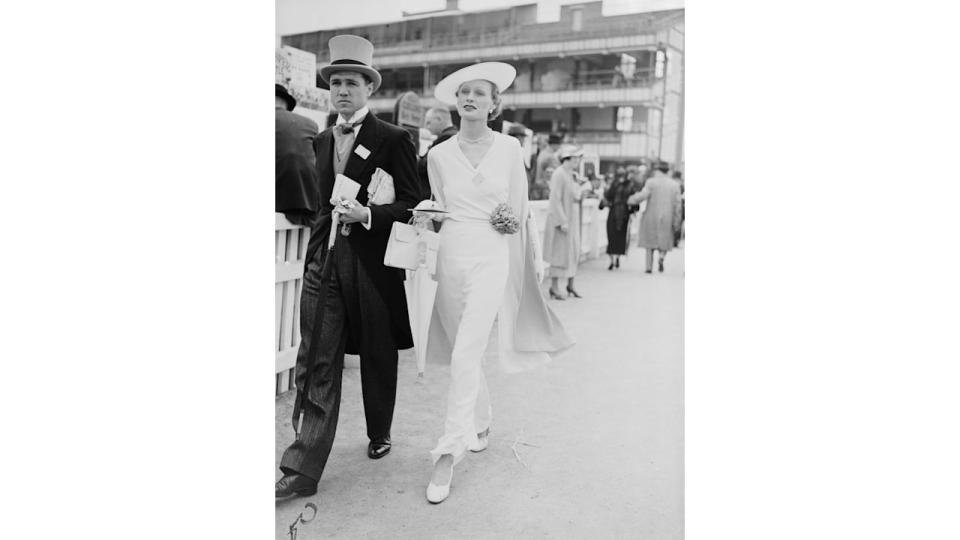 UNITED KINGDOM - NOVEMBER 07:  Couple at Royal Ascot, 1935. A photograph of a couple walking past racegoers at the Royal Hunt Cup horse-race, taken by Edward Malindine for the Daily Herald newspaper on 19 June, 1935. The woman appears in an elegant white ensemble, whilst her partner wears top hat and tails - traditional dress by the 1930s. The racecourse at Royal Ascot was founded by Queen Anne in 1711. As a racecourse favoured by British Royalty Ascot has become a highlight of the social season and is famous for its fashions, especially its hats. This photograph has been selected from the Daily Herald Archive, a collection of over three million photographs. The archive holds work of international, national and local importance by both staff and agency photographers.  (Photo by Daily Herald Archive/SSPL/Getty Images)