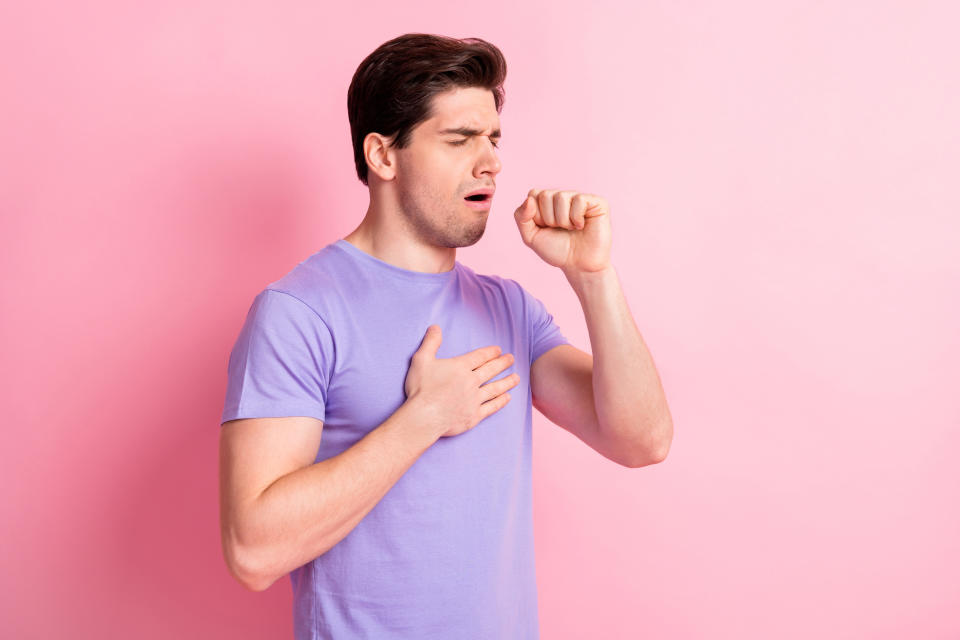 Man in purple T-shirt coughing into fist, appears to be demonstrating proper cough etiquette