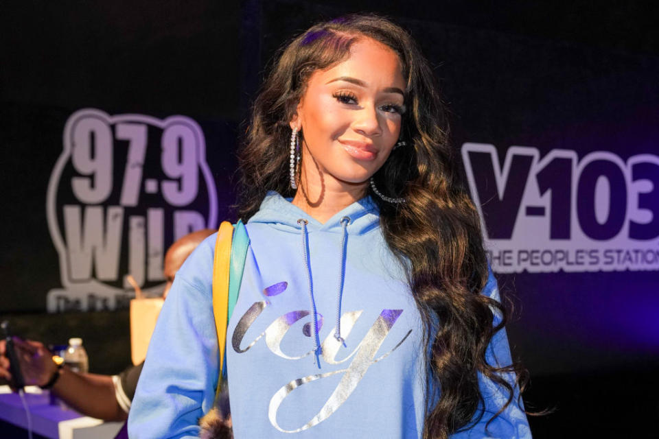 Saweetie at a V-103 event, smiling, wearing a casual "icy" hoodie and large hoop earrings, standing in front of a banner for 97.9 WILD