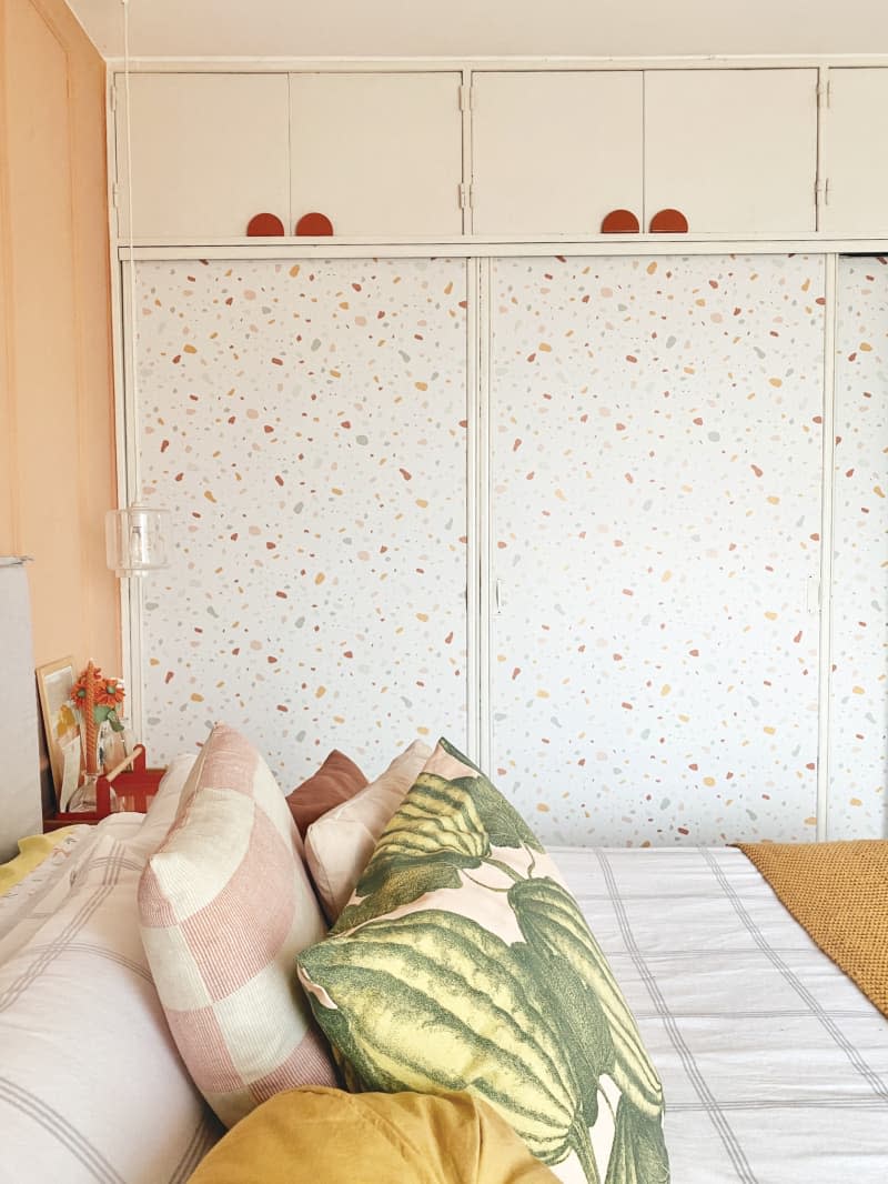 bedroom after repainting/remodel. Cantaloupe colored walls with decorative moulding, art prints of lamps, wood nightstand, white, ochre, blush, rose bedding with watermelon pattern pillow, cloest doors with terrazo pattern in white, gray, blush tones