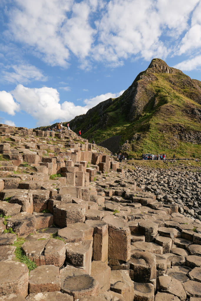 Giant's Causeway in Northern Ireland. Hexagonal basalt columns lead up a green hillside under a partly cloudy sky. Tourists are visible in the distance