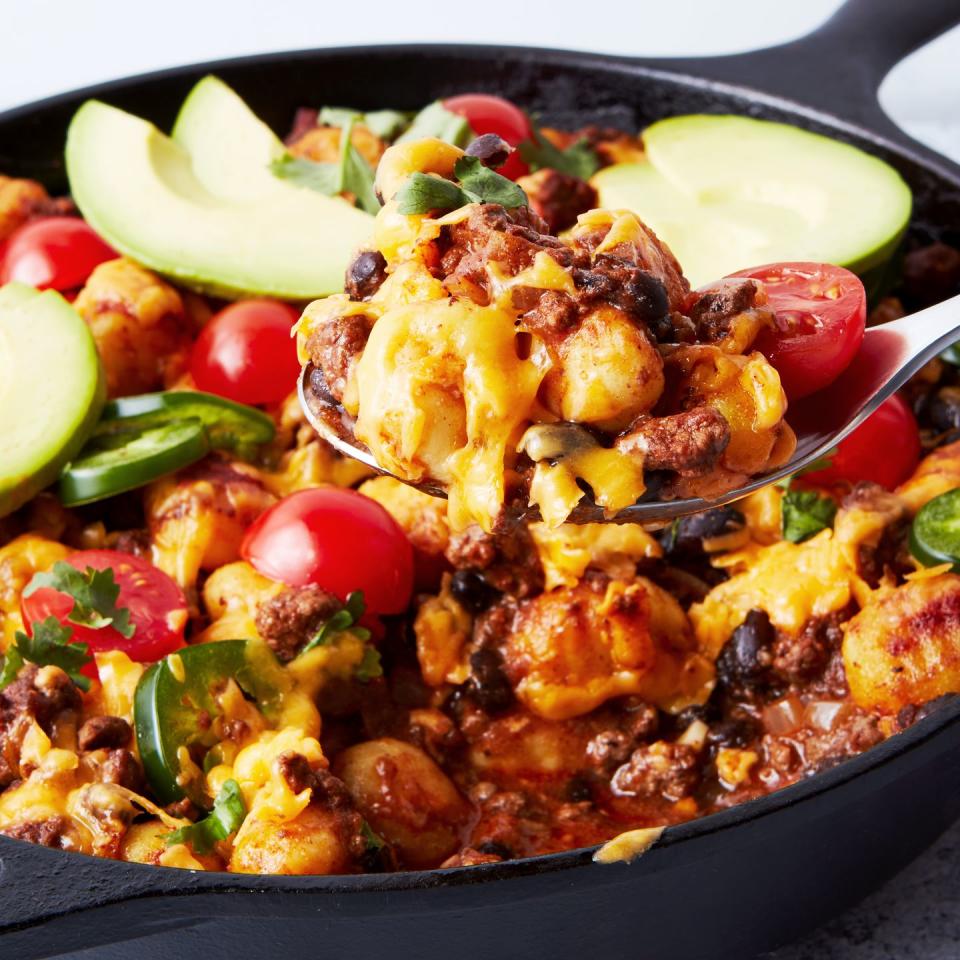 a pan with gnocchi covered in enchilada sauce, cheese, avocado, cilantro, and cherry tomatoes