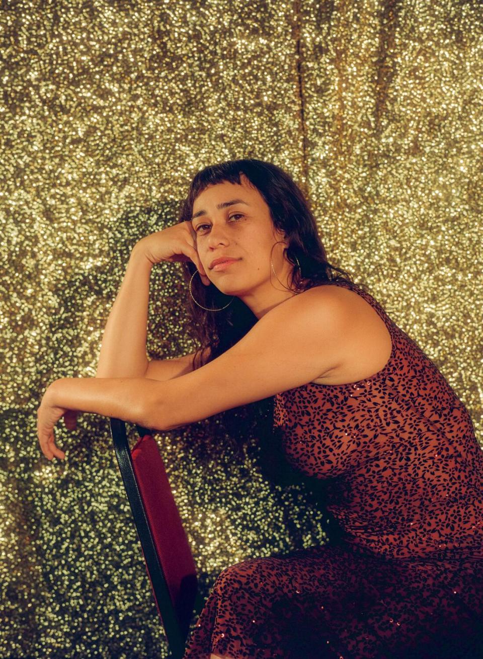 A woman leans against the back of a chair in front of a golden curtain.