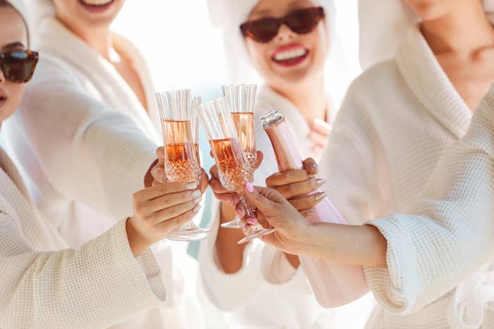 Four women in bathrobes toast with champagne glasses, each holding a glass of bubbly, one holding the bottle. They are smiling and wearing sunglasses