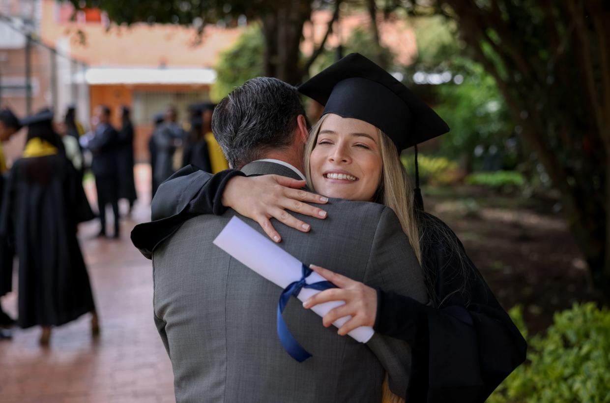 Happy graduate student hugging her father on graduation day while holding her diploma - education concepts