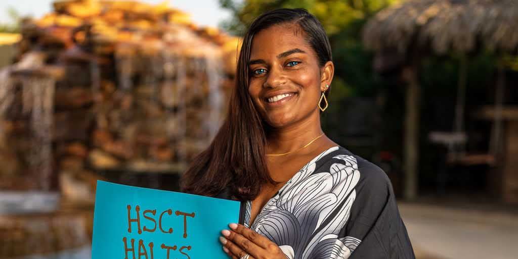 sagirah norris standing in front of a pool smiling wearing a long blue black and white dress, she carries a sign that lists her invisible disabilities