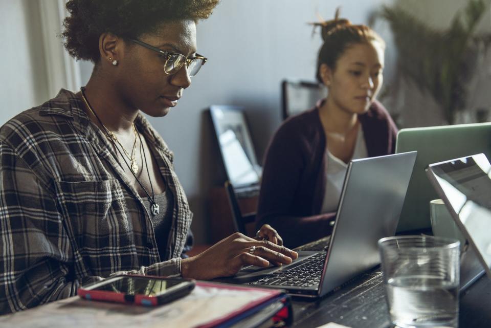 Two young women, one black and one Asian, sit together at a table while working on two laptops.