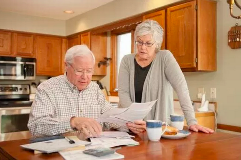 An older man and woman sitting at a kitchen table looking at paperwork.