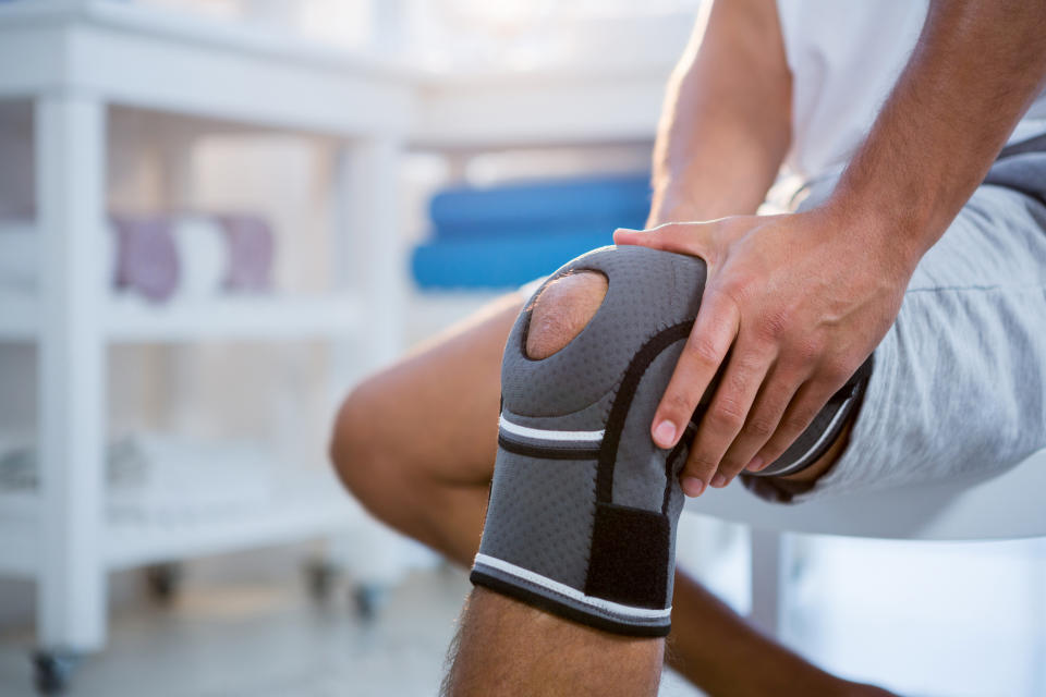 A person sits with a knee brace on their leg, holding the knee. The surrounding setting appears to be a clinic or medical office