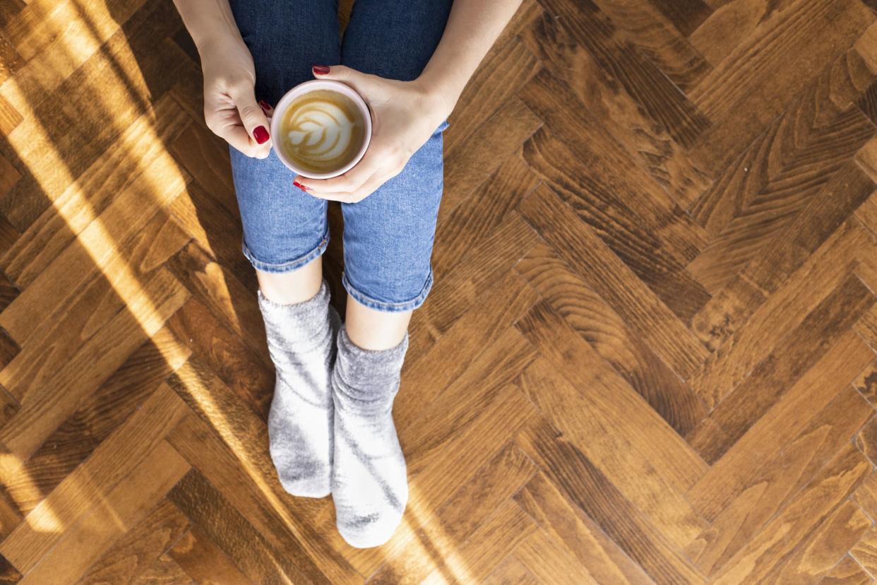 Woman's hands holding cup of coffee with a heart design in espresso, sitting on the wooden floor, showing jeans and grey socks, sunlight coming in from the left side