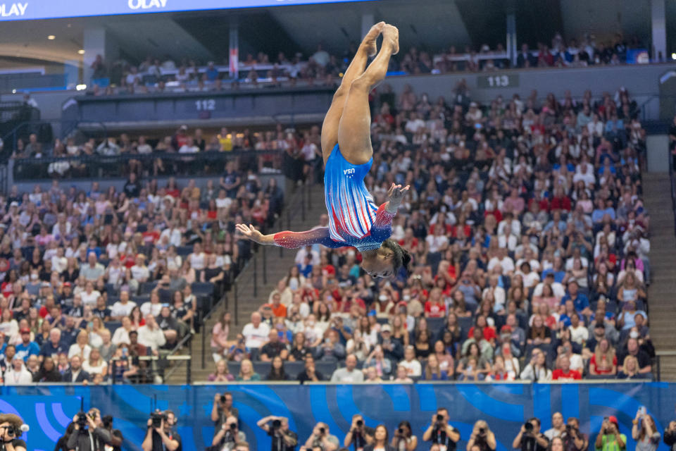 Simone Biles mid-air performing a gymnastics routine in front of a large audience. She is wearing a patriotic leotard with a blue and red design