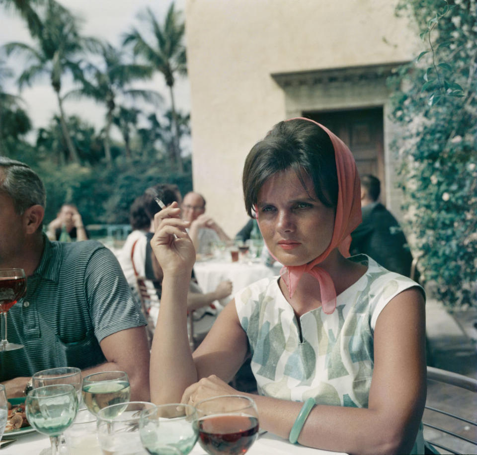 A woman in a patterned dress and headscarf sits at an outdoor table with drinks, holding a cigarette, surrounded by others dining