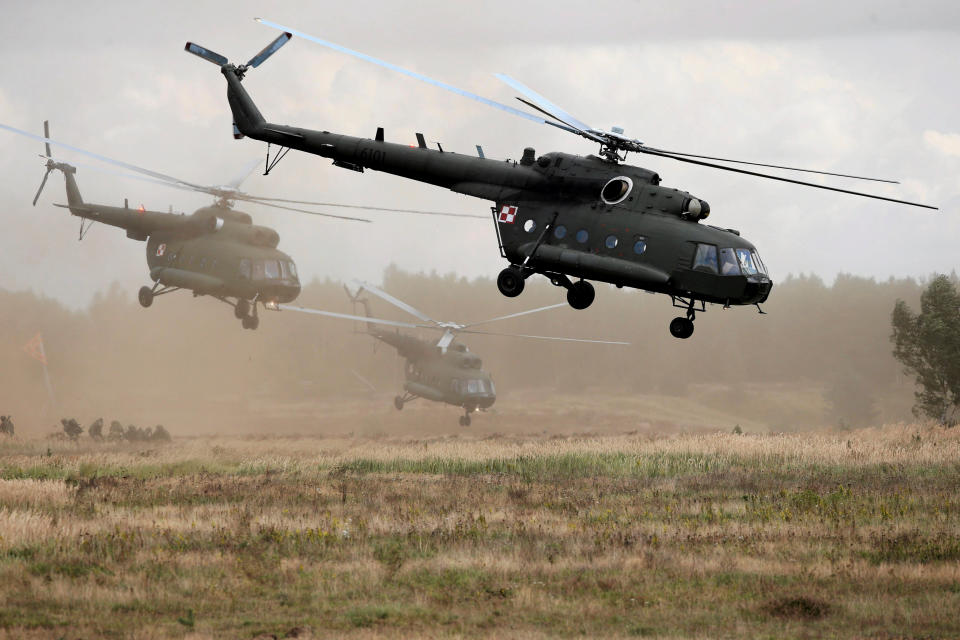 Three helicopters fly through mist above a tree-lined field with soldiers on the ground in the distance.