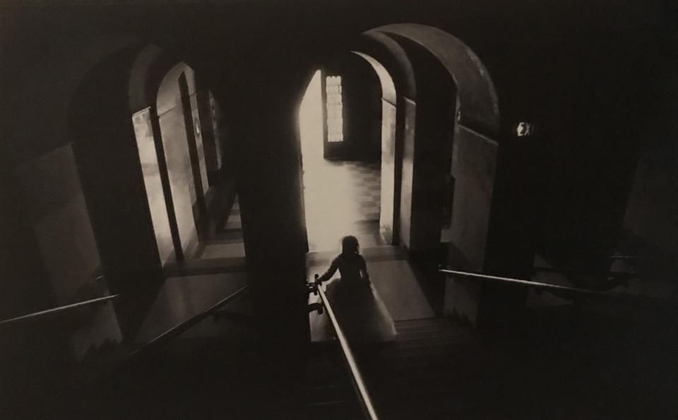 The silhouette of a girl walking down a shadowed staircase, one hand on the bannister.
