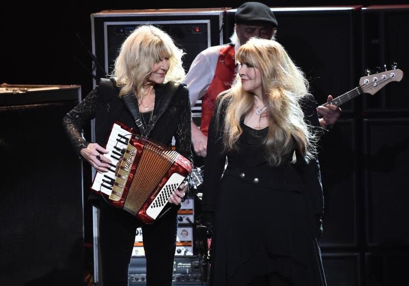 Two women with long blond hair in dark formal wear looking at each other while performing on a stage