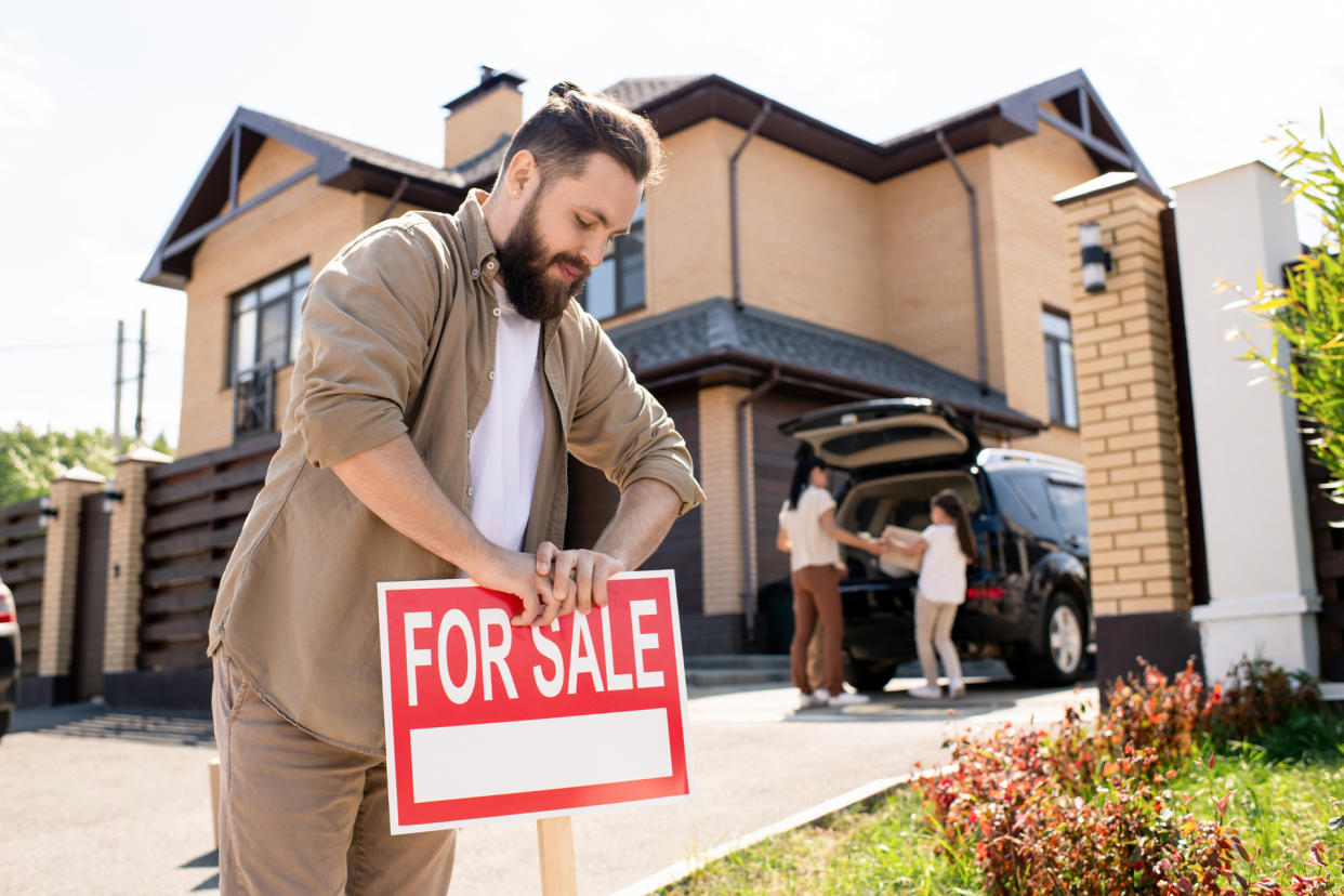 Young bearded father placing For sale sign into ground near house while putting house up for sale.