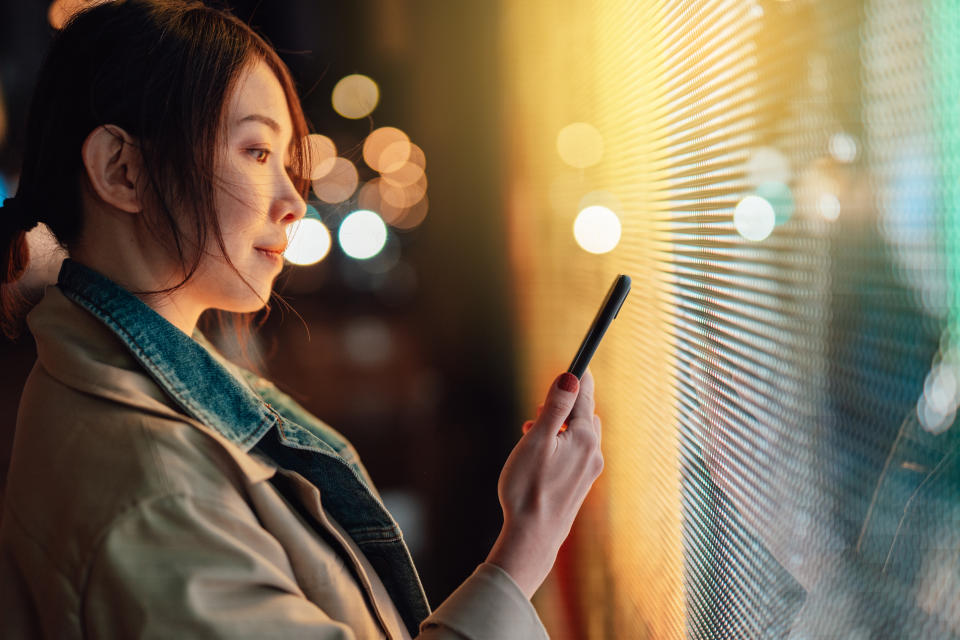 Young Asian woman using smartphone against illuminated digital display in city street at night. Blockchain technology. Trading cryptocurrency.  NFT (Non-Fungible Token) investment.