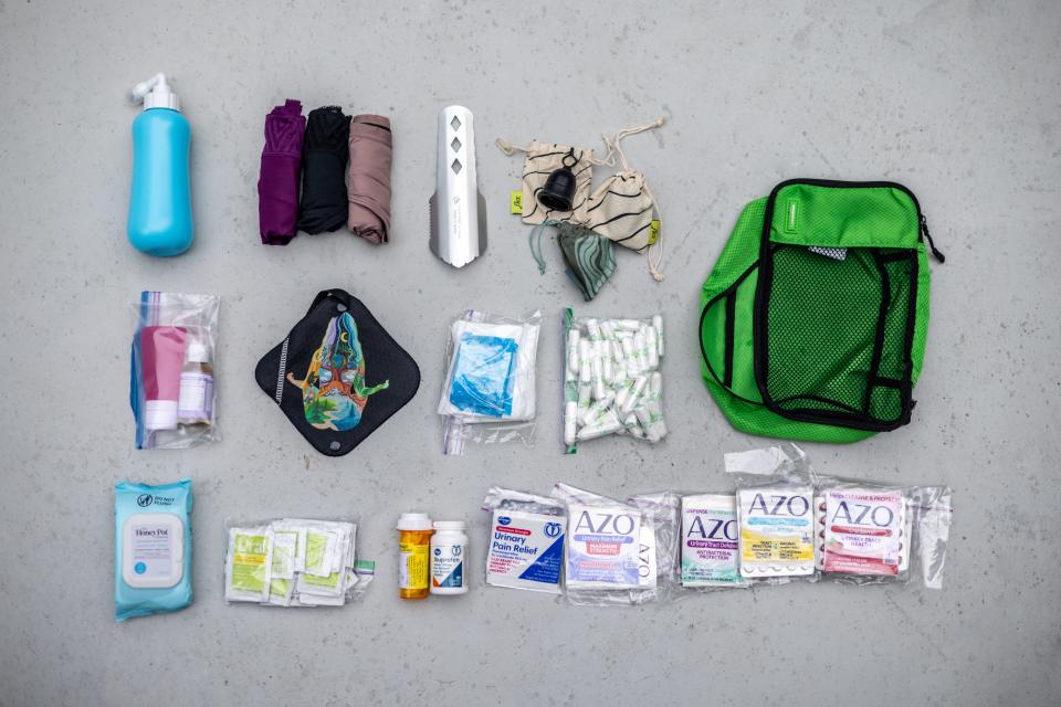 A photo from above of menstrual items in three rows. The first row has a field bidet, period underwear, field trowel, menstrual cups, and a green bag. The second has a Ziploc bag bag with a travel bottle and another unidentifiable bottle, black antimicrobial pee cloth, Ziploc bags with pads, and another Ziplock bag with tampons. The third row has wet wipes, Ural satchels, two bottles of antibiotics, then UTI medication. 
