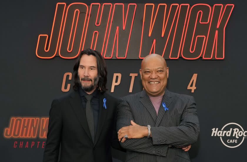 Two actors pose in front a movie poster that says 'John Wick'