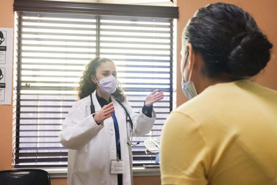 A masked woman in a white coat, left, gestures with her hands as she stands facing another woman