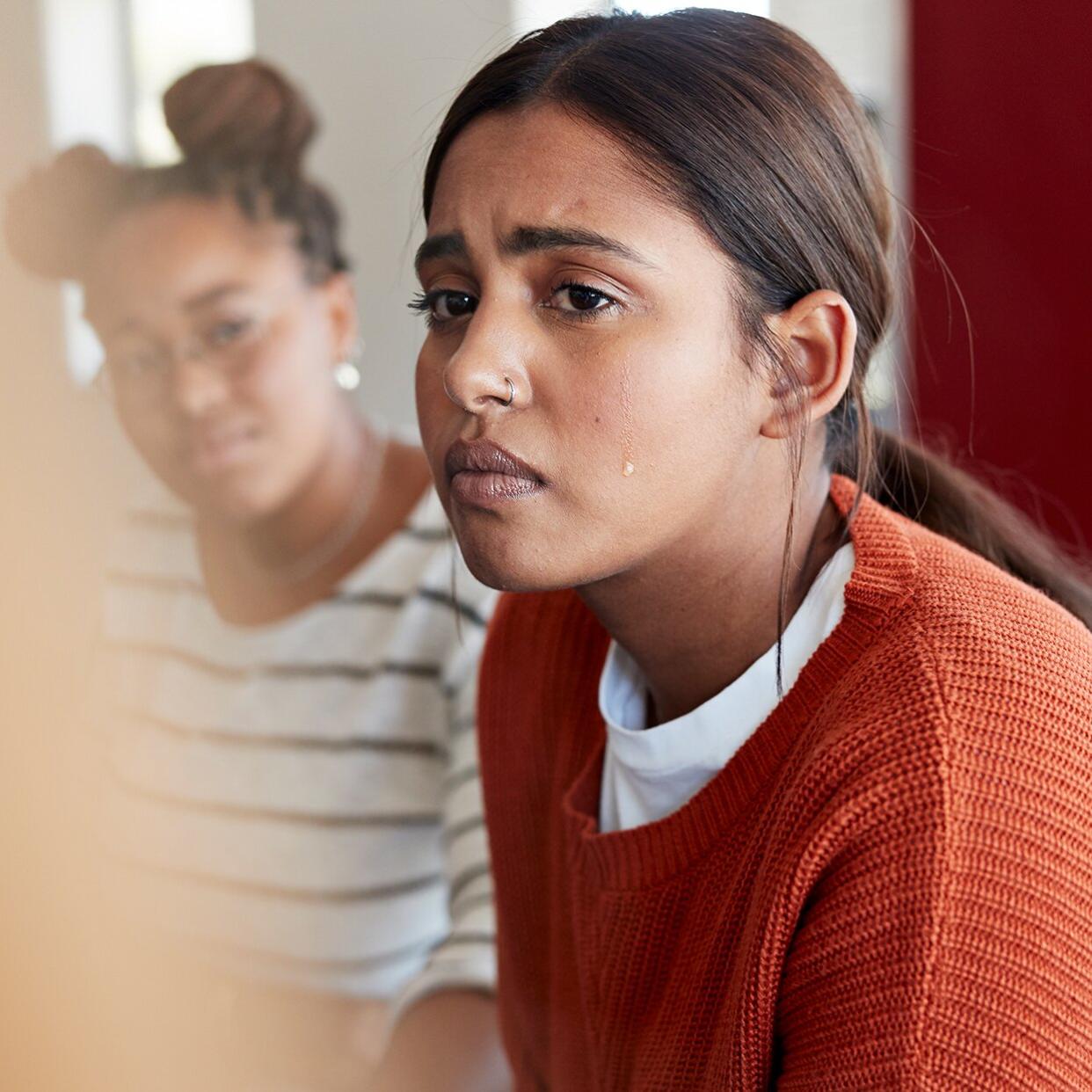 why am i crying for no reason?: woman furrowing her brows and crying, while a friend sits in the background looking sympathetic