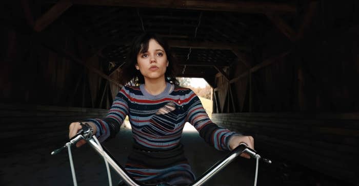 Jenna Ortega rides her bike across a covered bridge. She is wearing a striped sweater and looks ahead with a determined expression on her face.