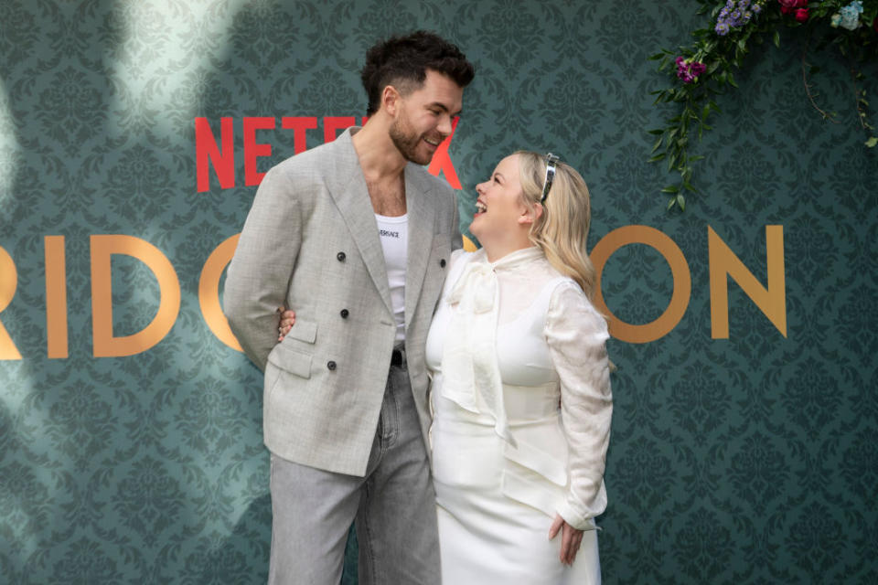 Luke Newton and Nicola Coughlan smiling at each other at a 'Bridgerton' event. Luke wears a light grey suit jacket and Nicola wears a white dress