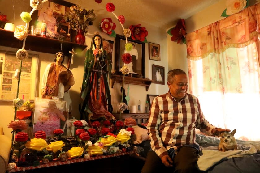 HUNTINGTON BEACH, CA - OCTOBER 11, 2020 - Hilario Herrera, 55, pets his dog Cuquis next to a statue of Our Lady of Guadalupe, that is used in religious celebrations, inside his apartment in the Oak View barrio in Huntington Beach on October 11, 2020. He lost his job as a soap factory worker and is struggling to get booked as a caterer. As a result, he doesn't know how he's going to fund a celebration to honor Mexico's patron saint, Our Lady of Guadalupe, on Dec. 12th. Herrera, who is known to throw over-the-top celebrations, is considering returning to his south-central Mexican state of Puebla. During this time of the year, preparations are underway for a handful religious and cultural celebrations put on by the community themselves. Instead residents wonder will there be any festivities to look forward to this year amid state mandates to avoid large gatherings and maintain social distance. (Genaro Molina / Los Angeles Times)