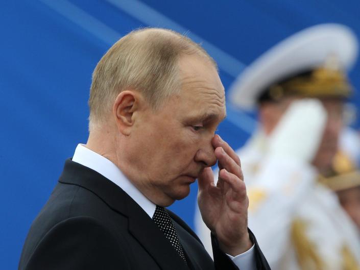 Russia president Vladimir Putin is shown looking down and touching his nose while military salute in the background.