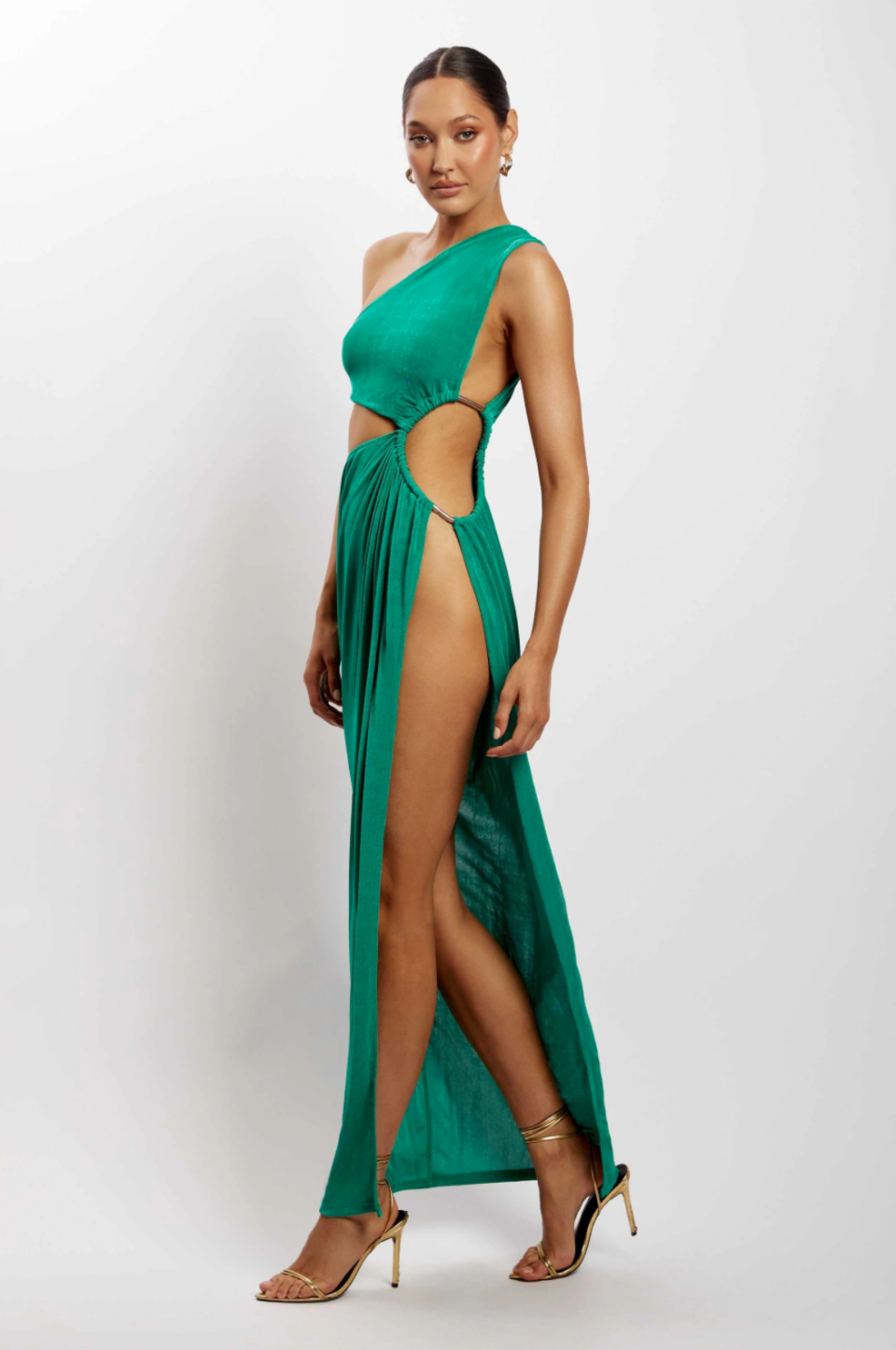 Woman with dark hair tied back wears a long green MESHKI PAIGE Ruched Side Cut Out Maxi Dress split on one side to the waist, with strappy high heels.