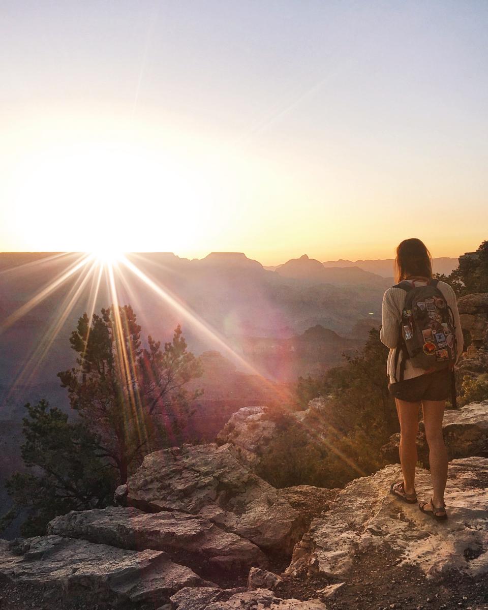 Emily, wearing sandals, shorts, a long-sleeved shirt, and a backpack, looks out over the sunrise on the mountains. 