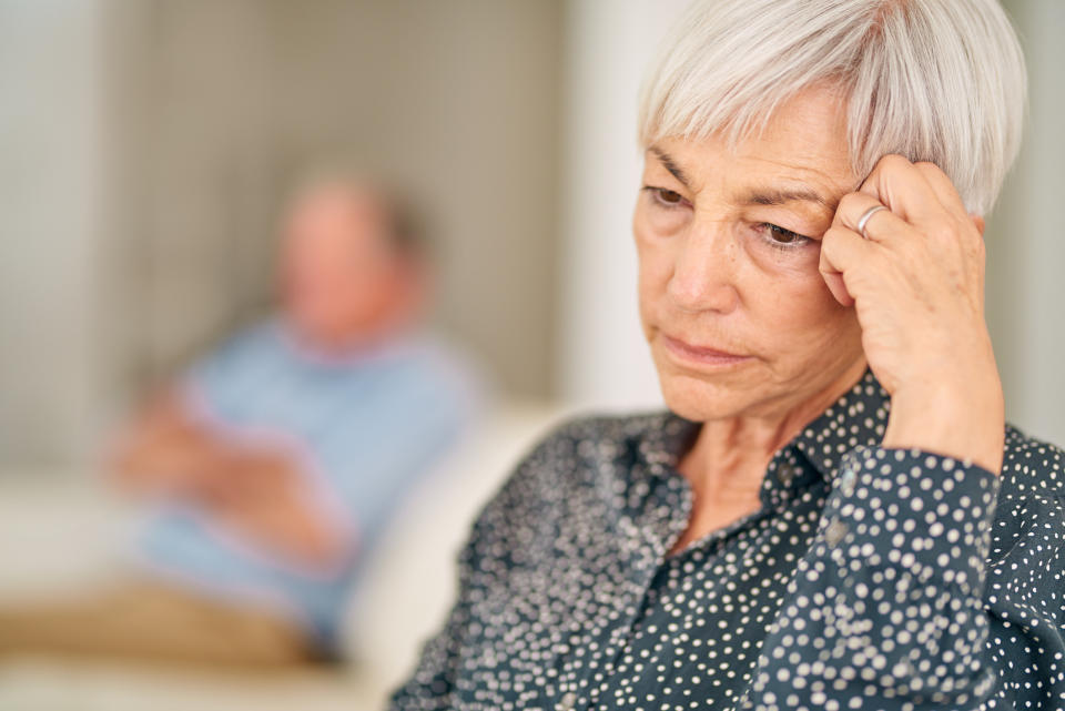 An older woman with short hair appears deep in thought while sitting, with a man blurred in the background