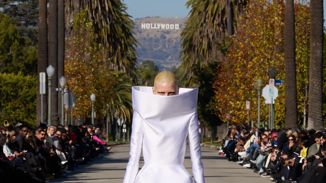 a model in balenciaga's fall 2024 show walks the outdoor runway in a white dress with a structured neckline so high it obscures her face
