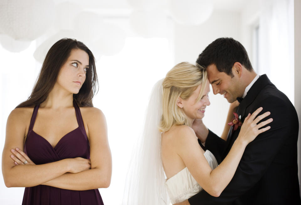 Woman in a bridesmaid dress looking jealous while a bride and groom embrace