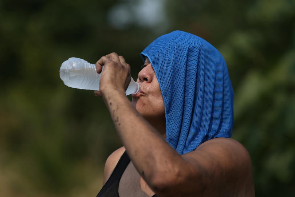 Cris drinks a bottle of water near her campsite, with a wet microfiber cloth draped over her head.