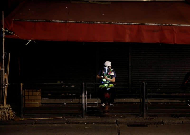 A journalist wearing a helmet and mask sits in the street while riot police prepare to disperse pro-democracy demonstrators gathering to commemorate the three-month anniversary of an assault by more than 100 men on protesters, commuters and journalists, in