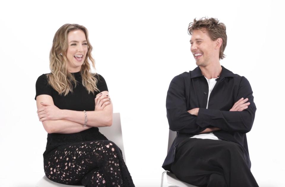 Jodie Comer and Austin Butler on chairs with crossed arms, laughing together. They are dressed in casual yet stylish clothing