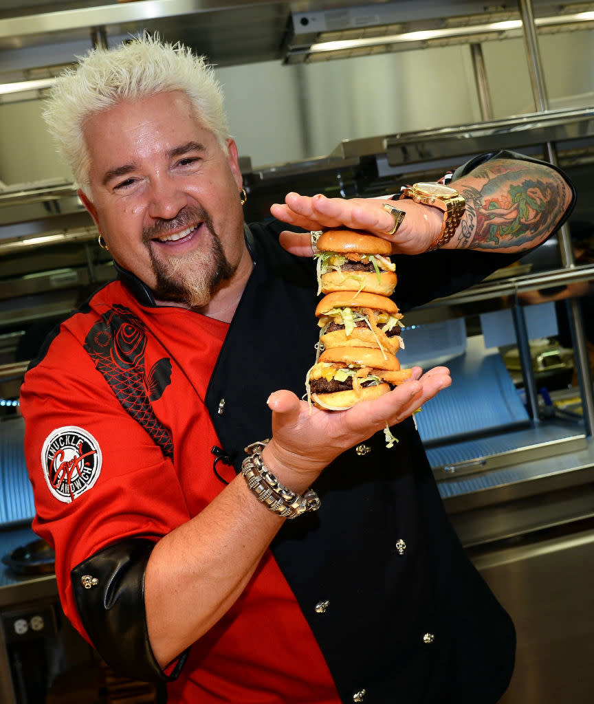 Guy Fieri in a chef's outfit, holding a tall stacked burger with multiple layers