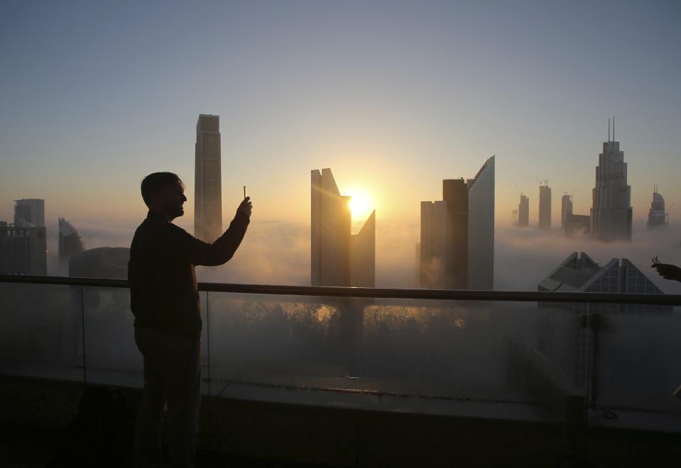 FILE- In this Saturday, Dec. 31, 2016 file photo, a man takes a photo as the sun rises over the city skyline from a balcony on the 42nd floor of a hotel on a foggy day in Dubai, United Arab Emirates. In an unmarked villa, nestled amid homes in an upscale Dubai neighborhood, sits the first fully functioning synagogue in the Arabian Peninsula in decades. Though its members keep its precise location secret, the synagogue’s existence and the tacit approval it has received from this Islamic sheikhdom represents a slow rebirth of a burgeoning Jewish community in the Persian Gulf uprooted over the decades after the creation of Israel. (AP Photo/Kamran Jebreili, File)