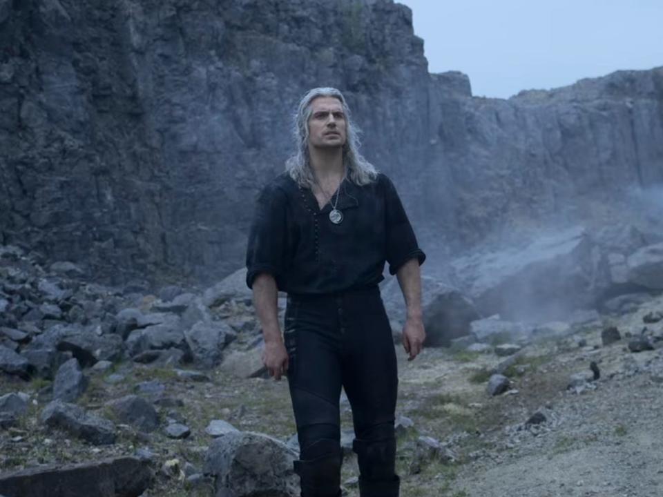 Henry Cavill with long white hair and a black outfit in front of mountains on "The Witcher"