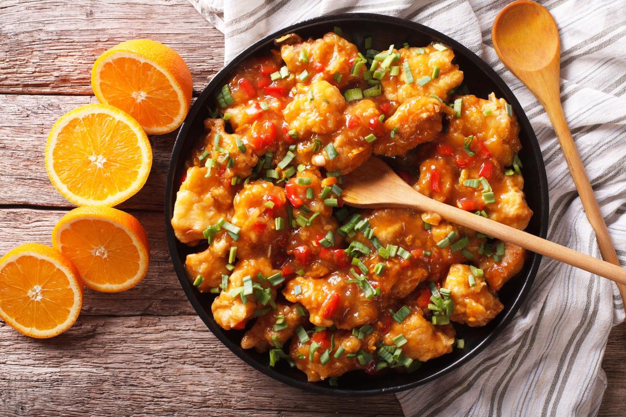 Spicy orange chicken stir-fry in a black ceramic bowl with a wooden spoon in it with four orange halves and another wooden spoon on the wide on a napkin on a rustic grey wooden table