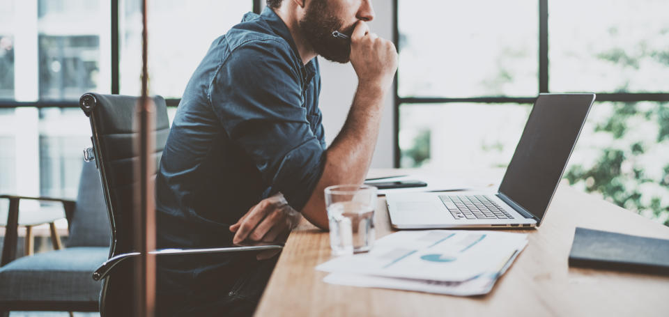 Young pensive coworker working at sunny work place loft while sitting at the wooden table.Man analyze document on laptop display.Blurred background.Horizontal wide