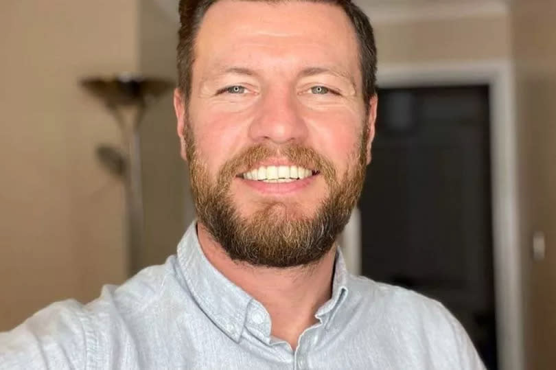 A man with brown hair, a quiff and a beard smiling