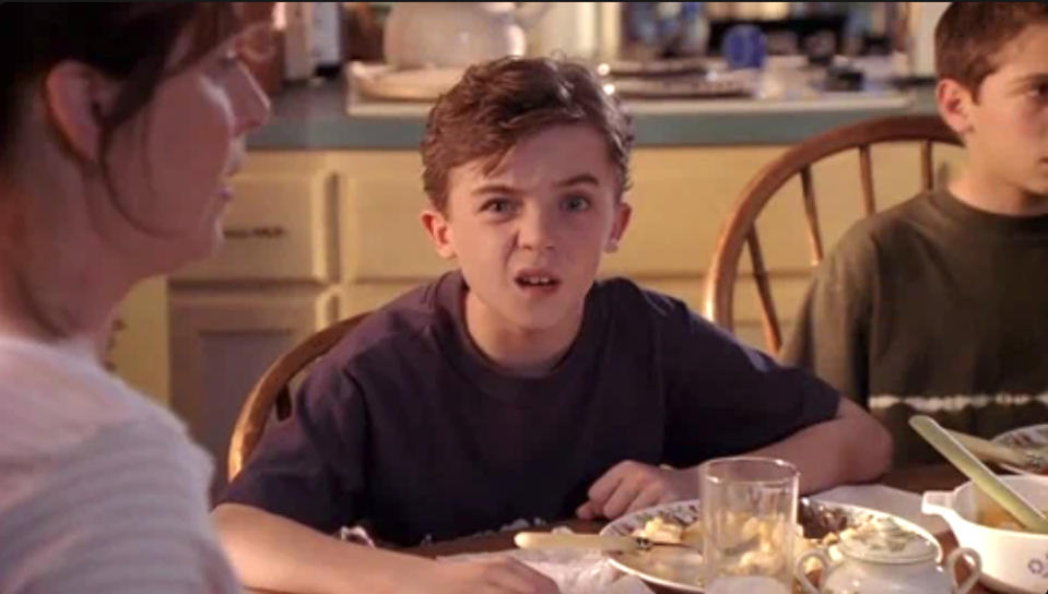 A confused boy sits at a dining table with an older woman and another boy. The boy has a puzzled expression, possibly reacting to something unexpected