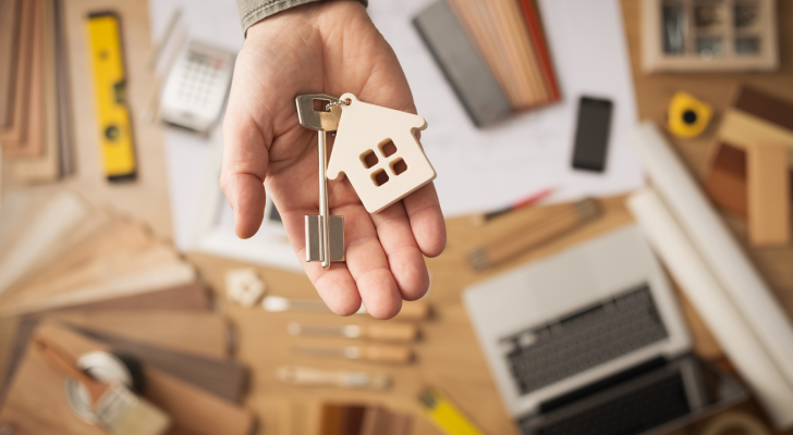 Real estate agent handing over a house key, desktop with tools, wood swatches and computer on background, top view. Real estate stocks.