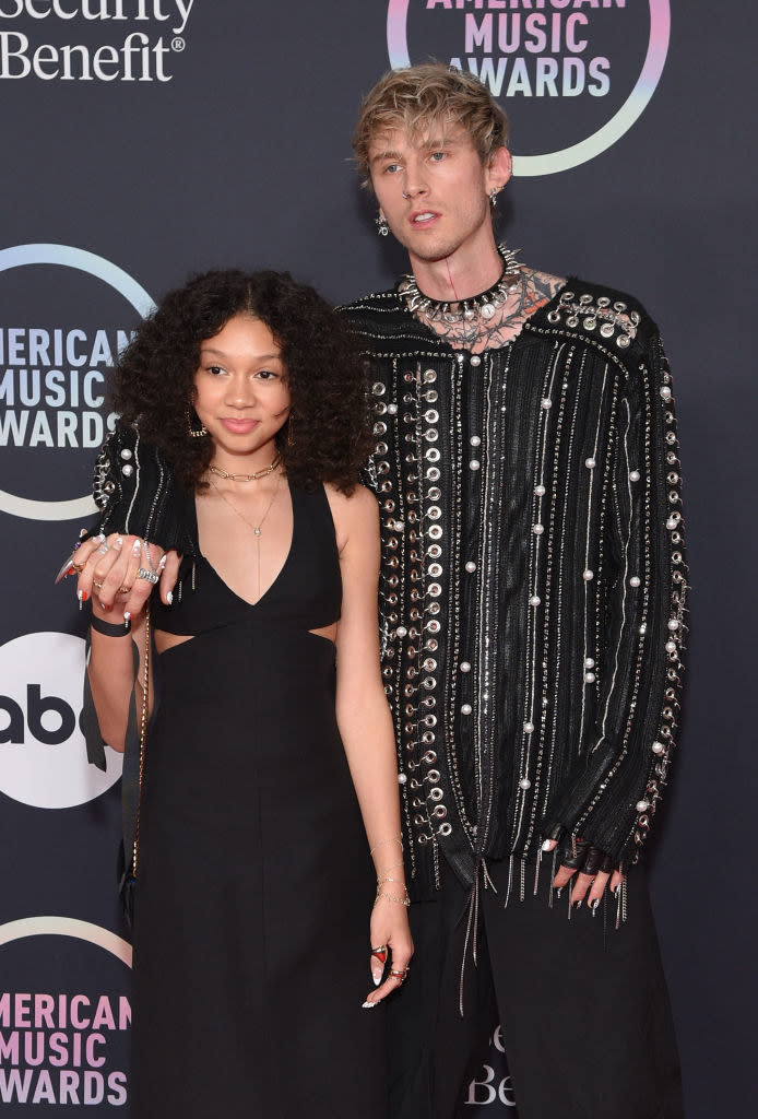 Casie Baker wears a thin strap dark gown and Machine Gun Kelly wears a studded long sleeve button up shirt